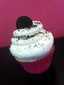 White cupcake with broken oreos folded into the batter, a vanilla buttercream topped with a mini oreo cookie and sprinkled with chocolate cookie crumbs.